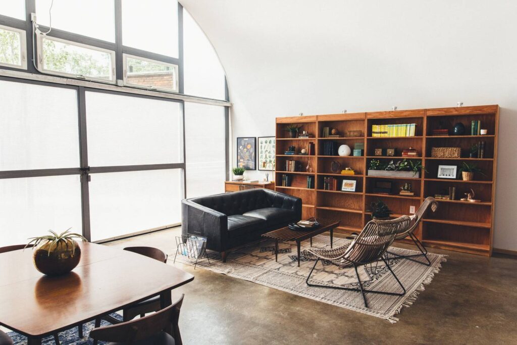 The living room of the QuonsetHaus with a black couch, coffee table and chairs and a large bookshelf with plants, books and other knick knacks.
