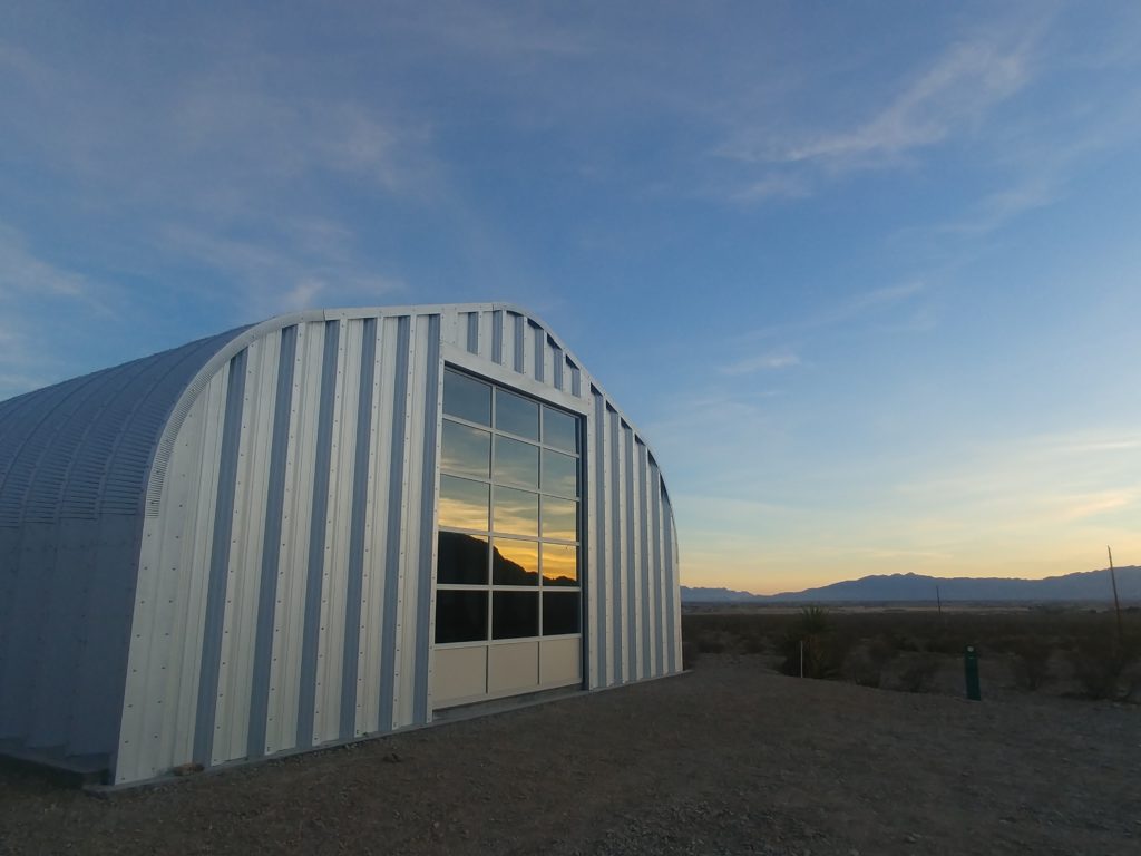 A-Model Quonset hut with steel end walls, glass garage door, a sunset reflecting off the garage door, and surrounded by deserts and mountains.