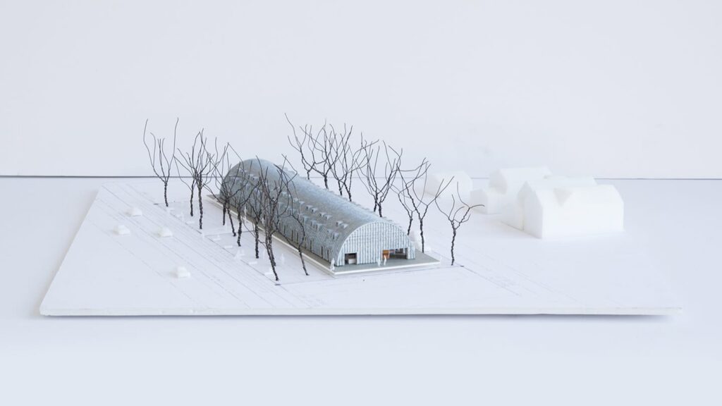 model of long steel arch building houses near by barren trees running along side