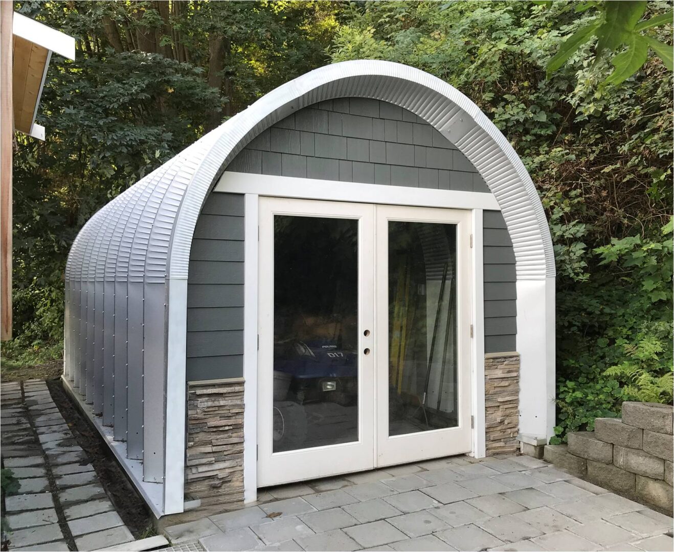 small s-model quonset hut in backyard with wood shingle and stone siding, french doors and atv inside