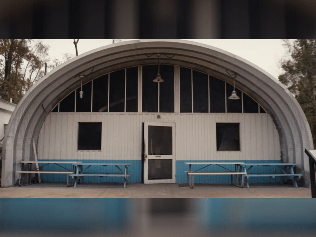 Steel S model quonset bbq pit house painted white and blue on front end wall. Has screen doors and screen windows on front endwall , with three lights hanging from the roof of quonset hut. Four picnic tables in front of building
