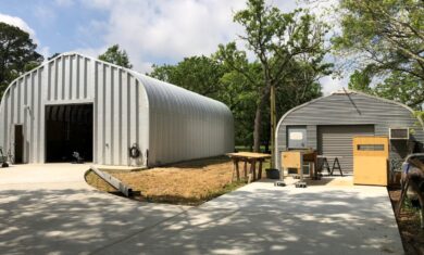 Driveways leading to two A-model Quonset huts: one with steel endwalls, large rolling door, front entrance; the other with custom ribbed grey endwall, matching rolling door, black entrance door, and workshop equipment in front.