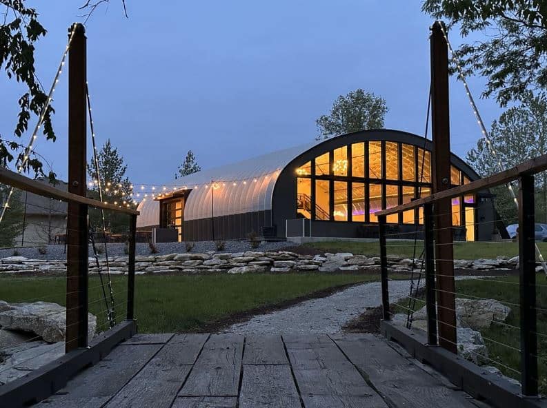 Wooden bridge with path leading to S-model Quonset hut with completely windowed endwall, building lit up by inside chandeliers, outside handing lights over side entrance to building