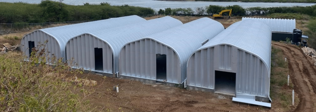 Four A-model Quonset huts attached side by side with front opening in metal endwalls, fifth quonset structure in the background, view of sea and other parts of the island in the background
