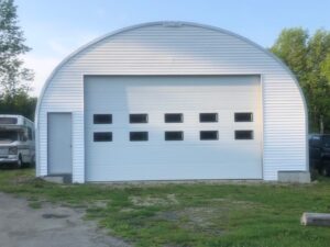 S-model quonset garage, custom white endwall, front entrance, white garage door, RV and trees to side