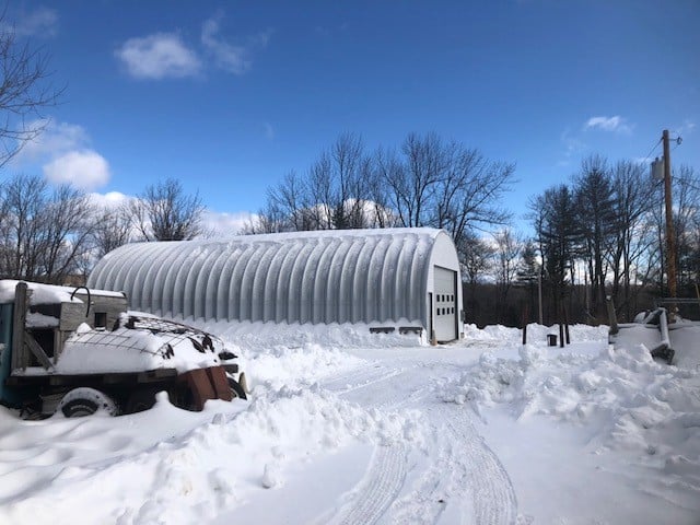 Side view of quonset with white garage door, snow on ground and on top of building