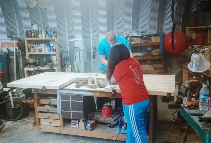 Inside Quonset workshop: two people working on wood project, various machinery and tools.