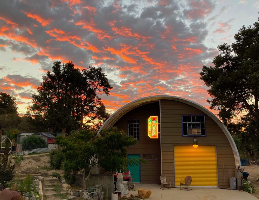 Q-model Quonset home with brown custom endwall partially recessed, overhead awning, lit neon sign, yellow rolling door, green entrance door, upper windows.