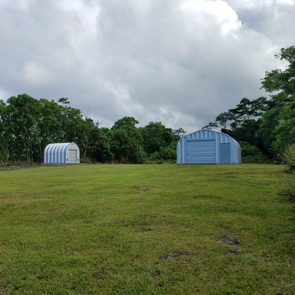 Two Quonset huts both with steel endwalls and rolling doors in Hawaiian landscape, larger A-model has front door entrance.