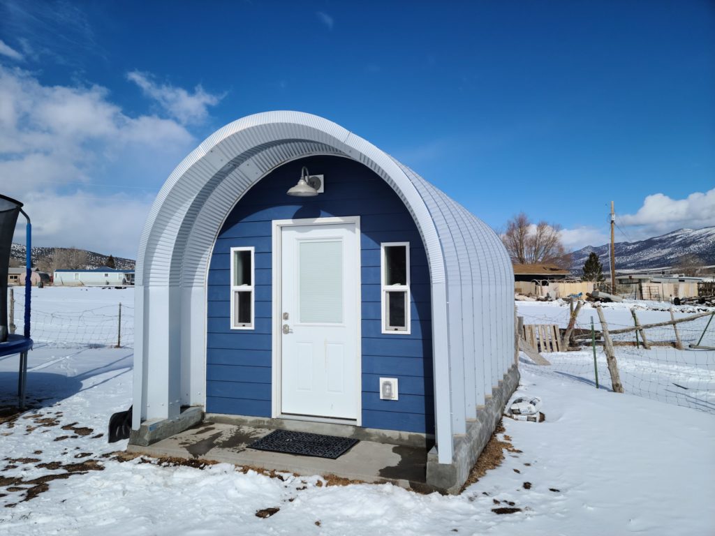 Tiny blue Quonset shed with custom endwall, white door, hanging entrance light, awning over porch, snowy lanscape.