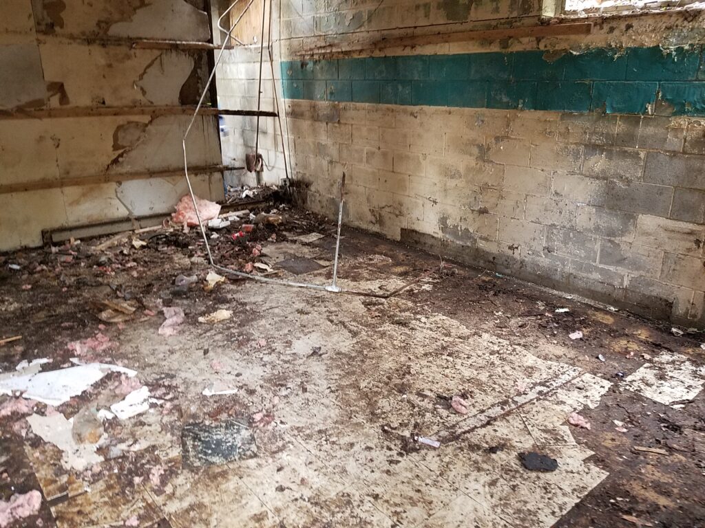 Inside crumbling abandoned building, dirt and trash on ground, hanging piping
