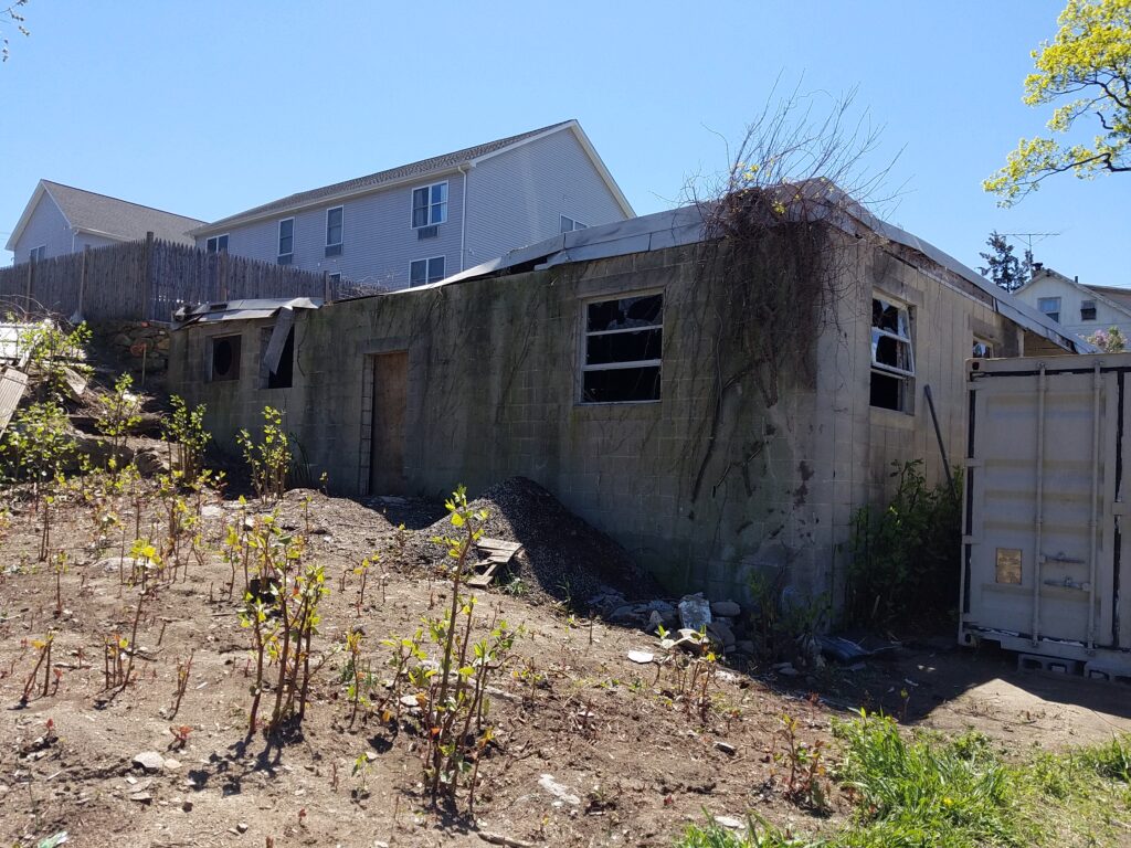 Abandoned building before restoration, grey bricks, plant growth on building, broken windows, dirt and plant overgrowth in front.