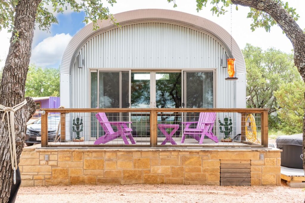 Front of Quonset airbnb on elevated stone platform, parch with purple chair seating area, sliding front doors, hummingbird feeder hanging from tree in front