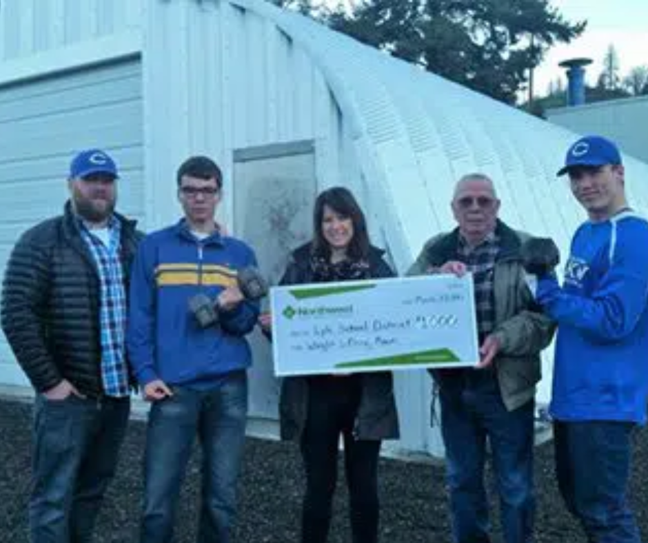 five people holding large donation check in front of quonset hut gym