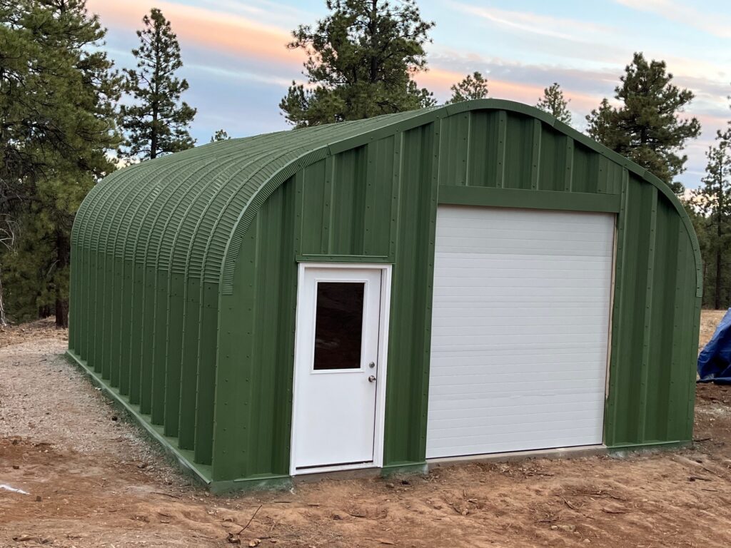 Green painted A-model Quonset hut, white front door, white rolling door, pine tree background