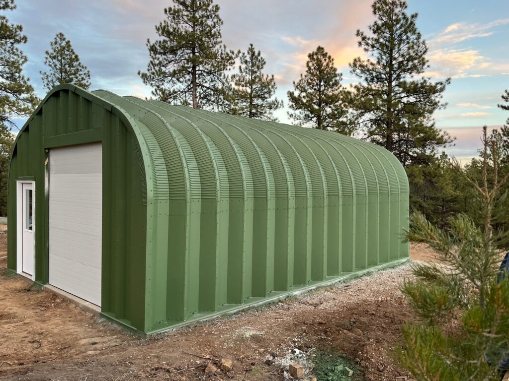 Green painted A-model Quonset hut, white front door, white rolling door, pine tree background