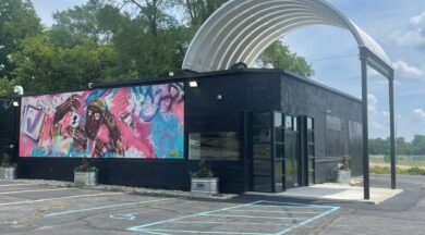 A steel arched roofing system covering a portion of a small black building with a pink and blue mural painted on the side