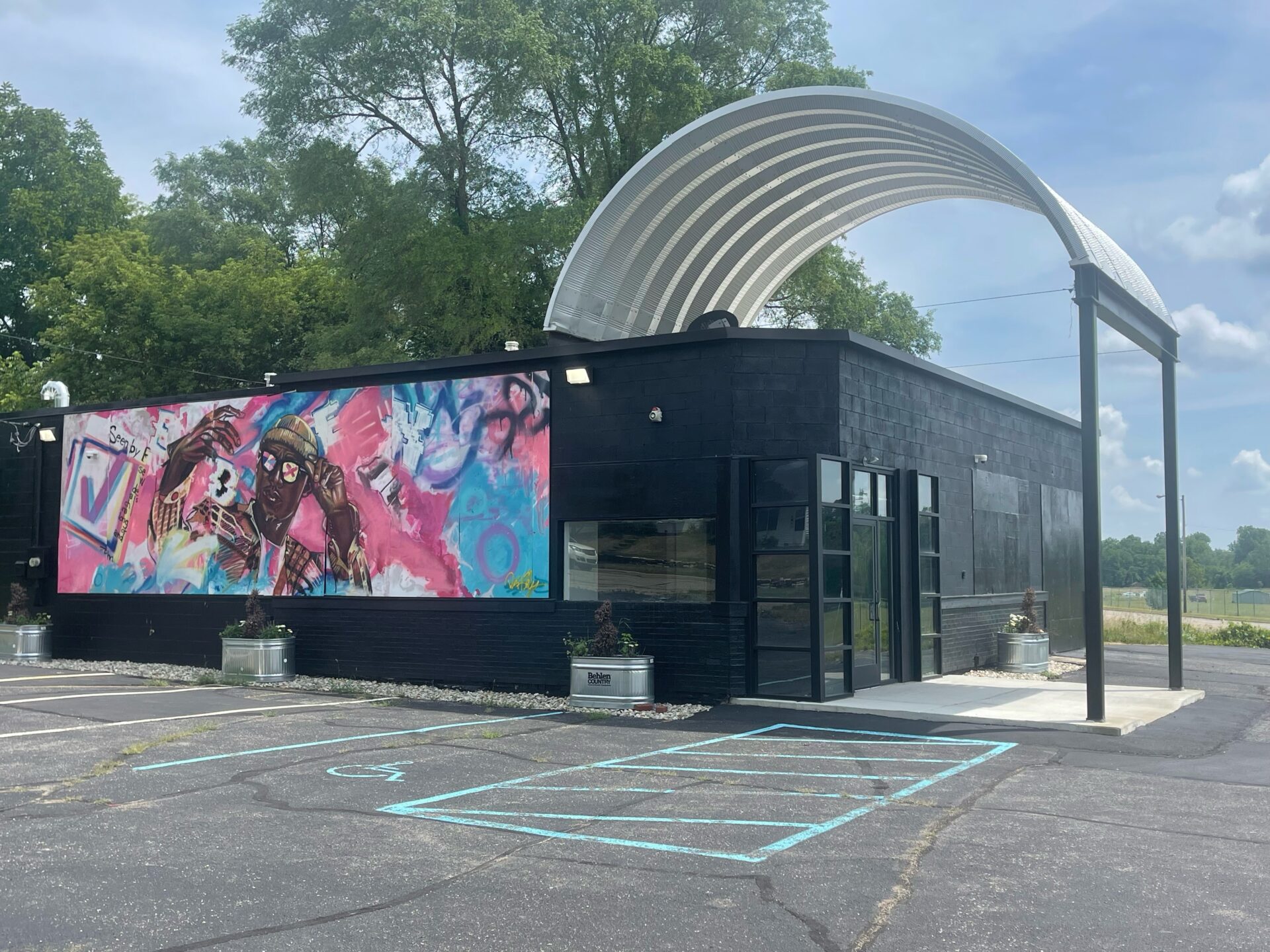 A steel arched roofing system covering a portion of a small black building with a pink and blue mural painted on the side