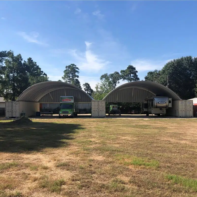 Shipping container storage with three freight containers and two metal arches; camper, truck, and other equipment sheltered underneath.