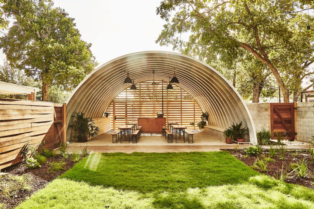 Open-ended event space covered by metal arches, overhead hanging lights, planked back endwall with spaces, picnic tables, and bar.