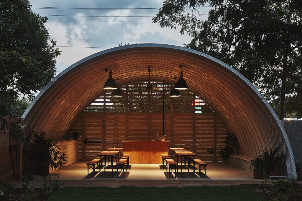 Open-ended event space covered by metal arches, overhead hanging lights, planked back endwall with spaces, picnic tables, and bar.