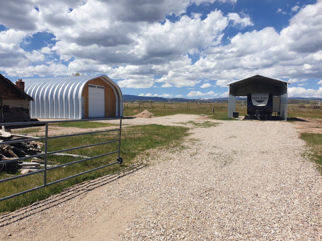 A-Model Quonset hut with custom wood end wall. white garage door, next to a car cover with an RV underneath it.