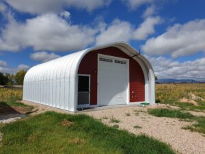 A-Model Quonset hut with custom red end wall, white garage door, man door, a green hose on the bottom right of the building, and mountains in the background.