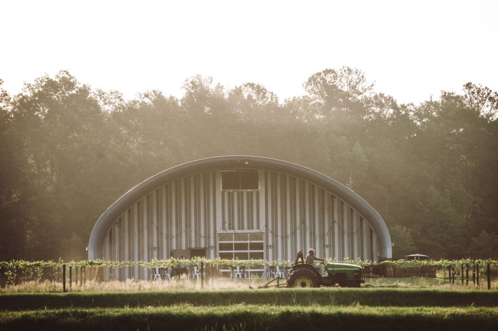 Open ended S-model pavilion, suspended lights above seating area, crops and man on tracker plowing soil in front.