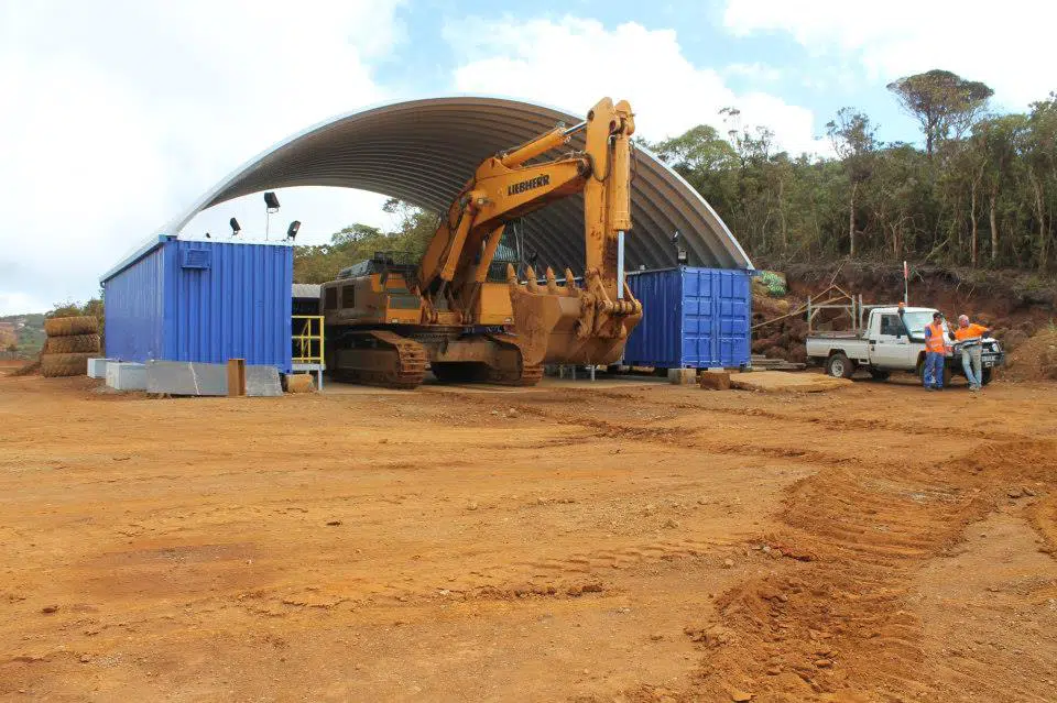 Metal arch roof system set on top of blue freight containers sheltering a large Liebherr excavator at a construction site, two construction workers by a white truck are set near the container cover.