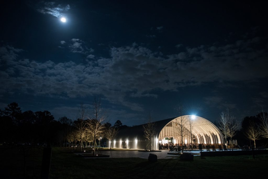 Night view of lit S-model food pavilion, suspended lights, and people at picnic style dining tables.