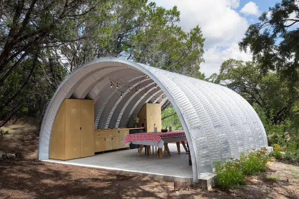 Open-ended Quonset pavilion on cement foundation with built in kitchen, suspended overhead lighting, and picnic table.