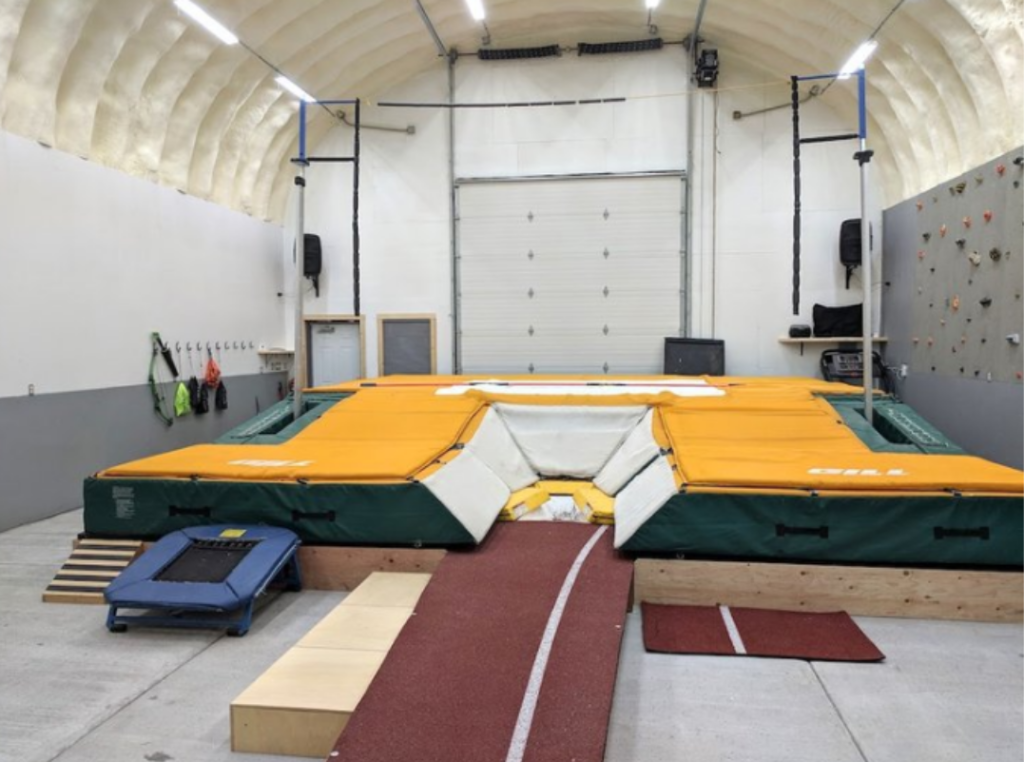 Inside a custom built Quonset hut: insulated main room with equipment for pole vaulting, a rock climbing wall, an entrance door, and an insulated garage door.