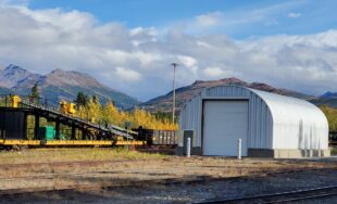 S-Model Quonset hut with two white poles in front, a ramp going up on the left, and two mountains in the background with clouds.