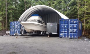 Container cover on top of two blue freight container, small boat underneath, pine trees behind steel structure