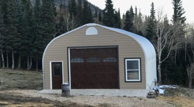 A-Model Quonset hut with tan end wall, brown garage and man door, a window to the right, and a stump in front.