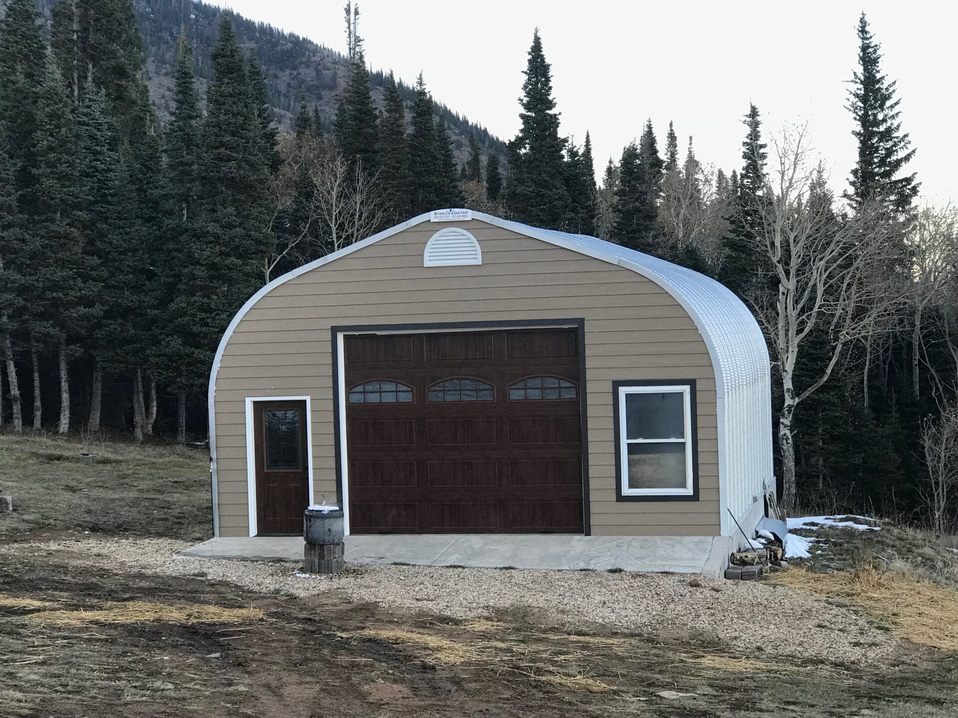 A-Model Quonset hut with tan end wall, brown garage and man door, a window to the right, and a stump in front.