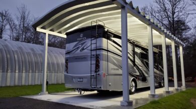 Large grey and black RV parked under a steel carport lit up at night