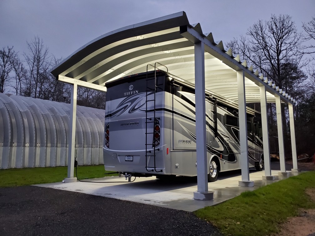 Large grey and black RV parked under a steel carport lit up at night