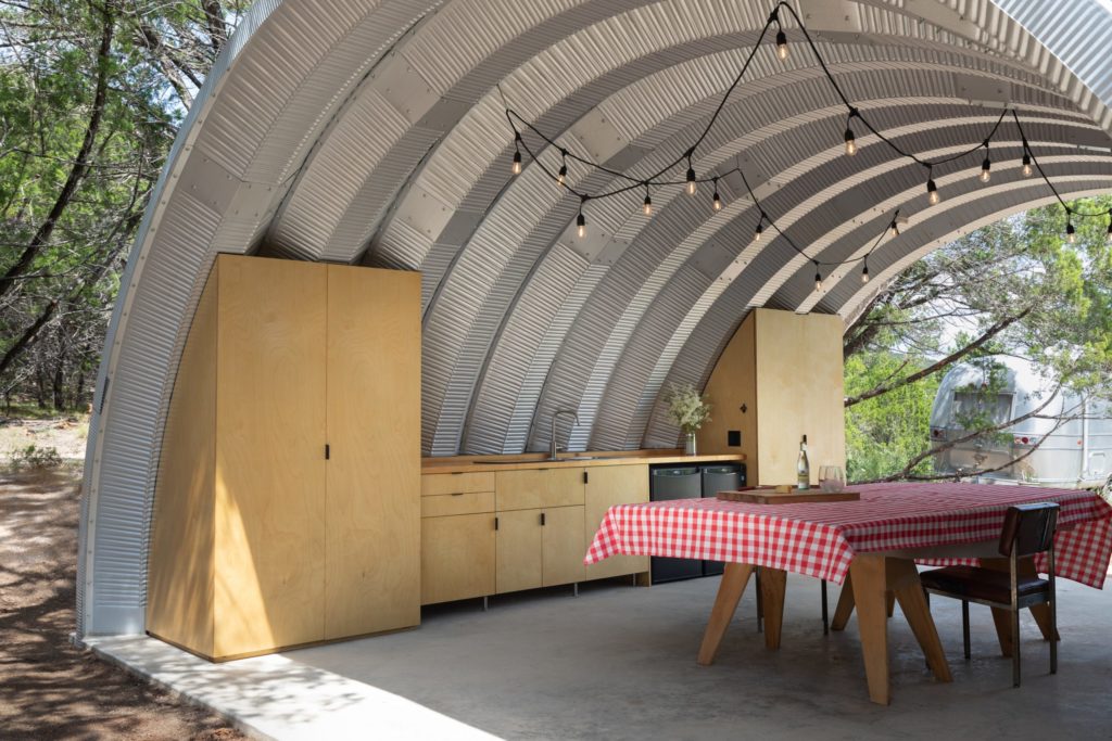 Inside open-ended Quonset pavilion on cement foundation with built in kitchen, suspended overhead lighting, and picnic table.