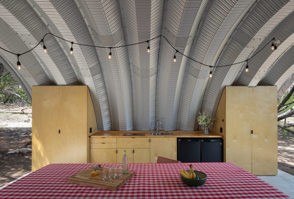 Inside open-ended Quonset pavilion with built in kitchen, suspended overhead lighting, and picnic table.