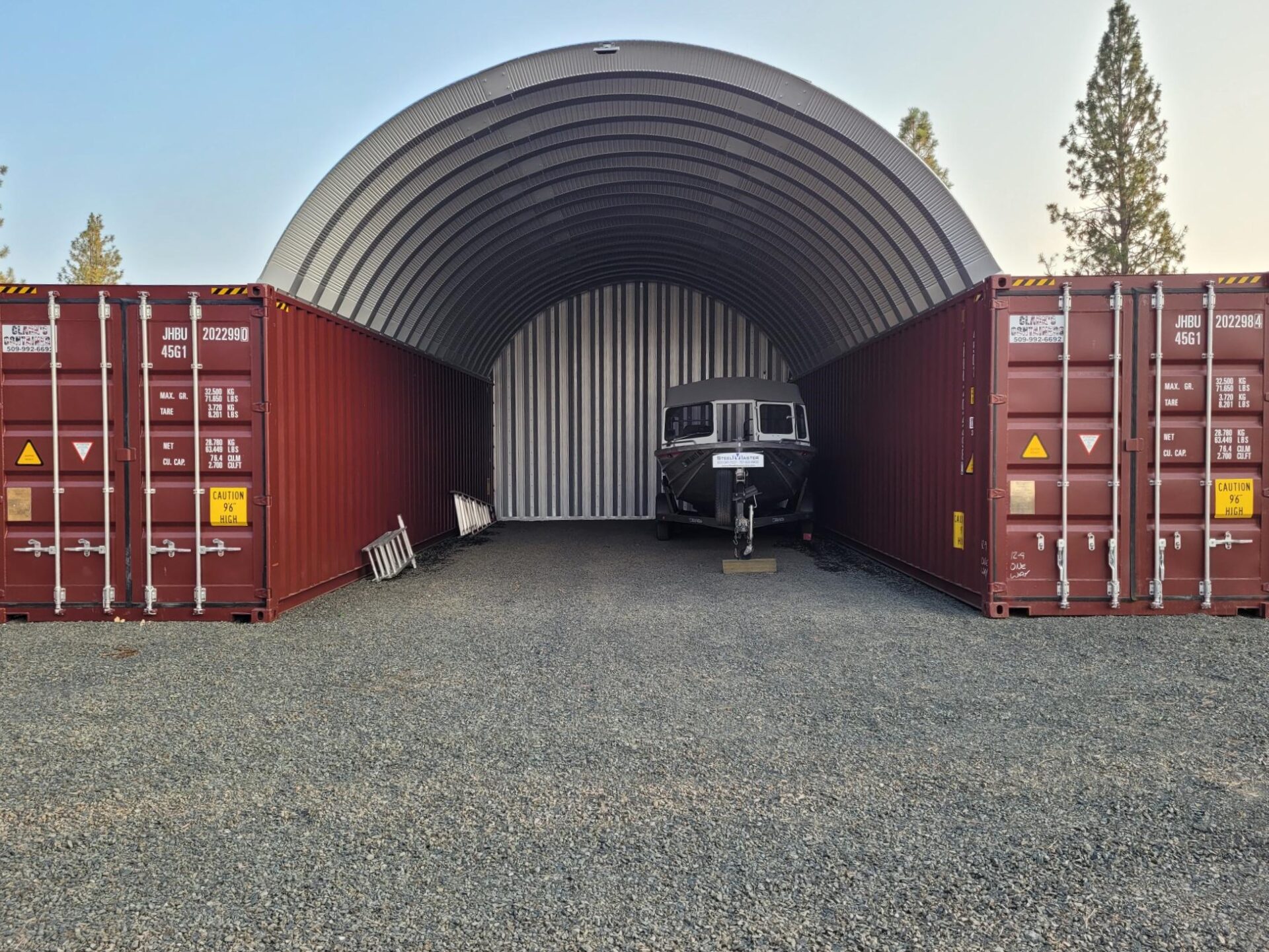 steel arch roof mounted on two red shipping containers with solid rear steel endwall and a boat inside of the structure