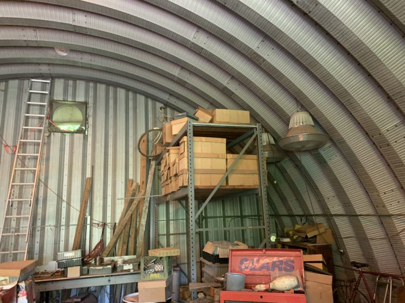 Inside Quonset workshop with metal endwall, shelving and various stored items.