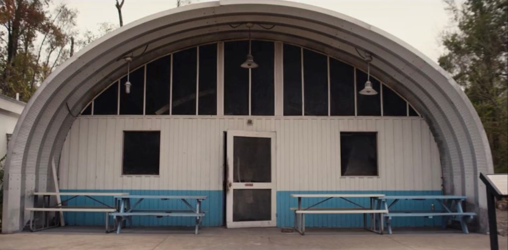 S-model Quonset Pit House with extended awning over blue and white picnic benches, hanging overhead lights, custom white endwall with low blue stripe, windows, and large center door ajar.