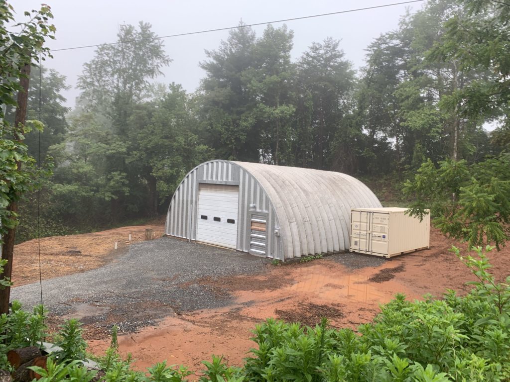 Q-model Quonset workwork in woods, metal endwall, white garage door, tan shipping container to side of building.