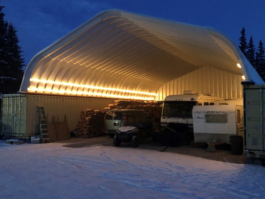 Night photo of container cover with back steel endwall, open structure has side strip lighting and shows storage of wood, van, RV, camper, and equipment, outside ground covered in snow.