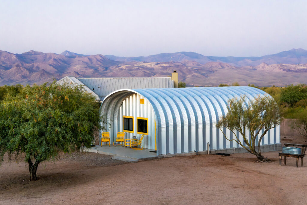 Far view of S-Model Quonset hut with three yellow chairs, two windows bordered in yellow, with a mountain range and house in the background.