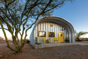 S-Model Quonset hut in the daytime with steel end walls, a tree to the left, three yellow chairs, and a yellow man door with a black symbol on it.