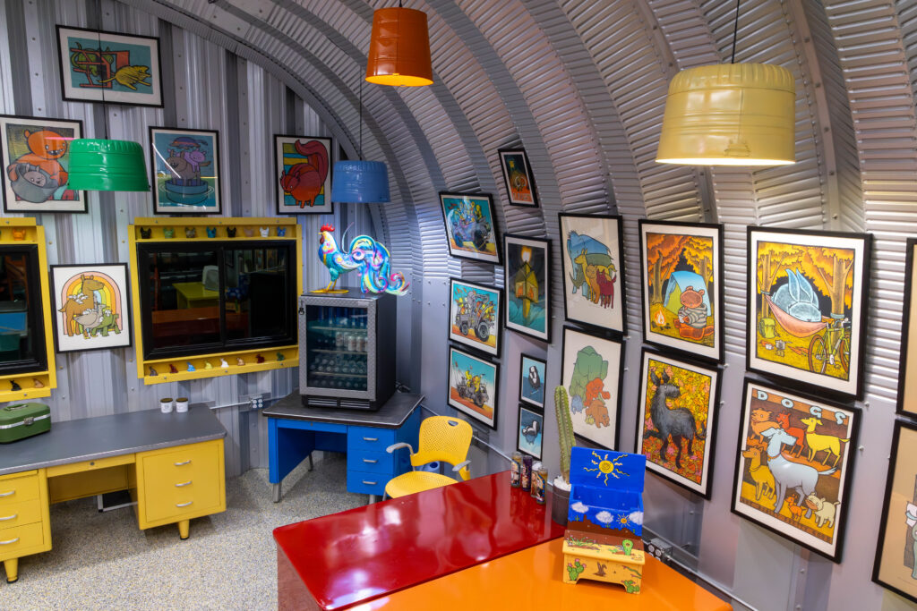 Inside of an S-Model Quonset hut with multiple paintings, a glass chicken on top of a mini fridge, and four multi-colored hanging lights.