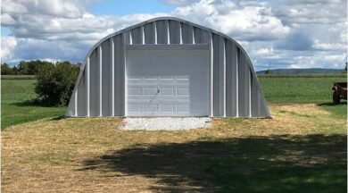 X-Model Quonset hut with steel end wall. bush on the left. and part of a trailer on wheels on the right.