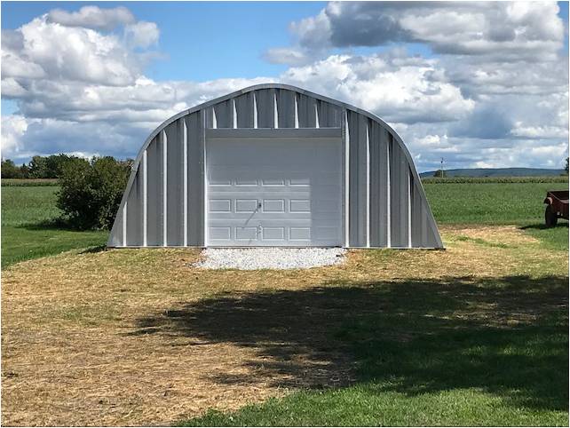 X-Model Quonset hut with steel end wall. bush on the left. and part of a trailer on wheels on the right.